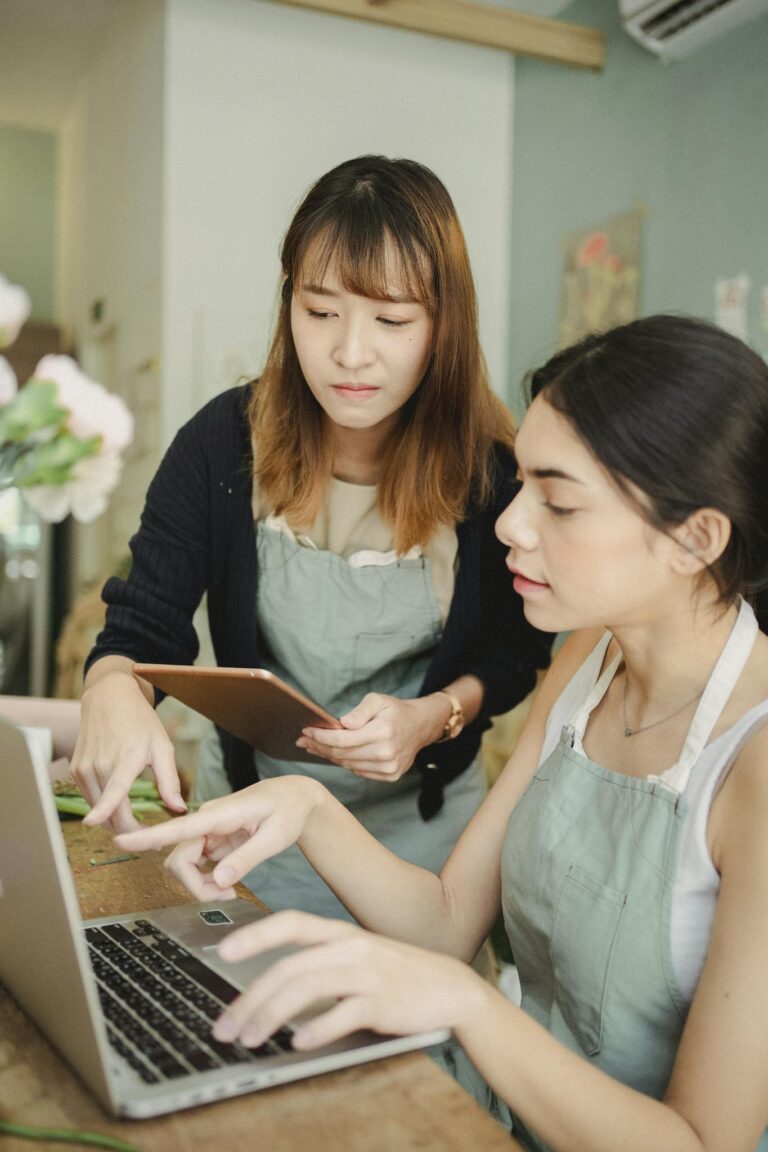 Focused multiethnic female colleagues in floral store at table working on computer while checking orders in internet during work in floristry studio