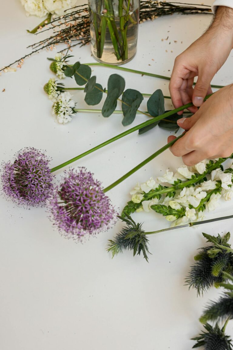 Hands arranging various flowers on a white table, showcasing a creative floral arrangement process.